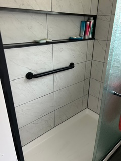 Shower with white tile, black handrail, and recessed shelf holding toiletries.