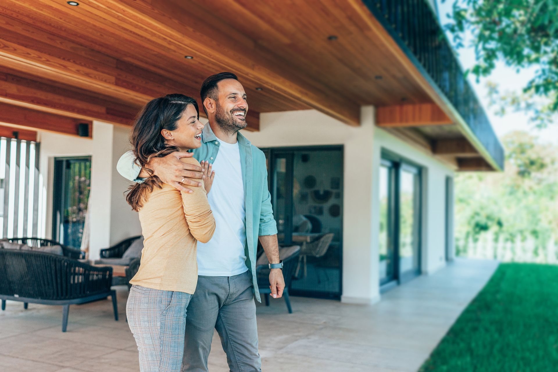 A couple stands on a modern patio with a wood-paneled ceiling, smiling and looking toward their home.