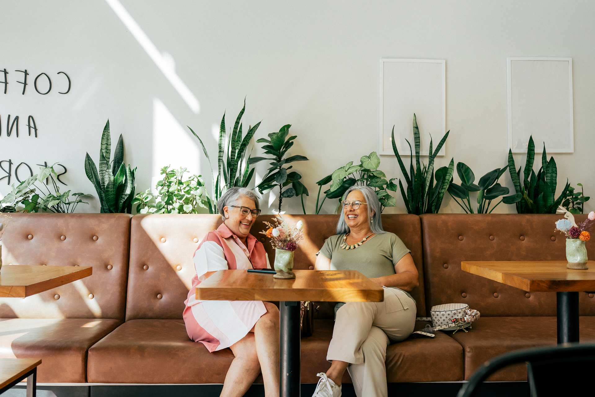 Two people smiling and conversing while sitting at a table in a modern cafe with many indoor plants behind them.