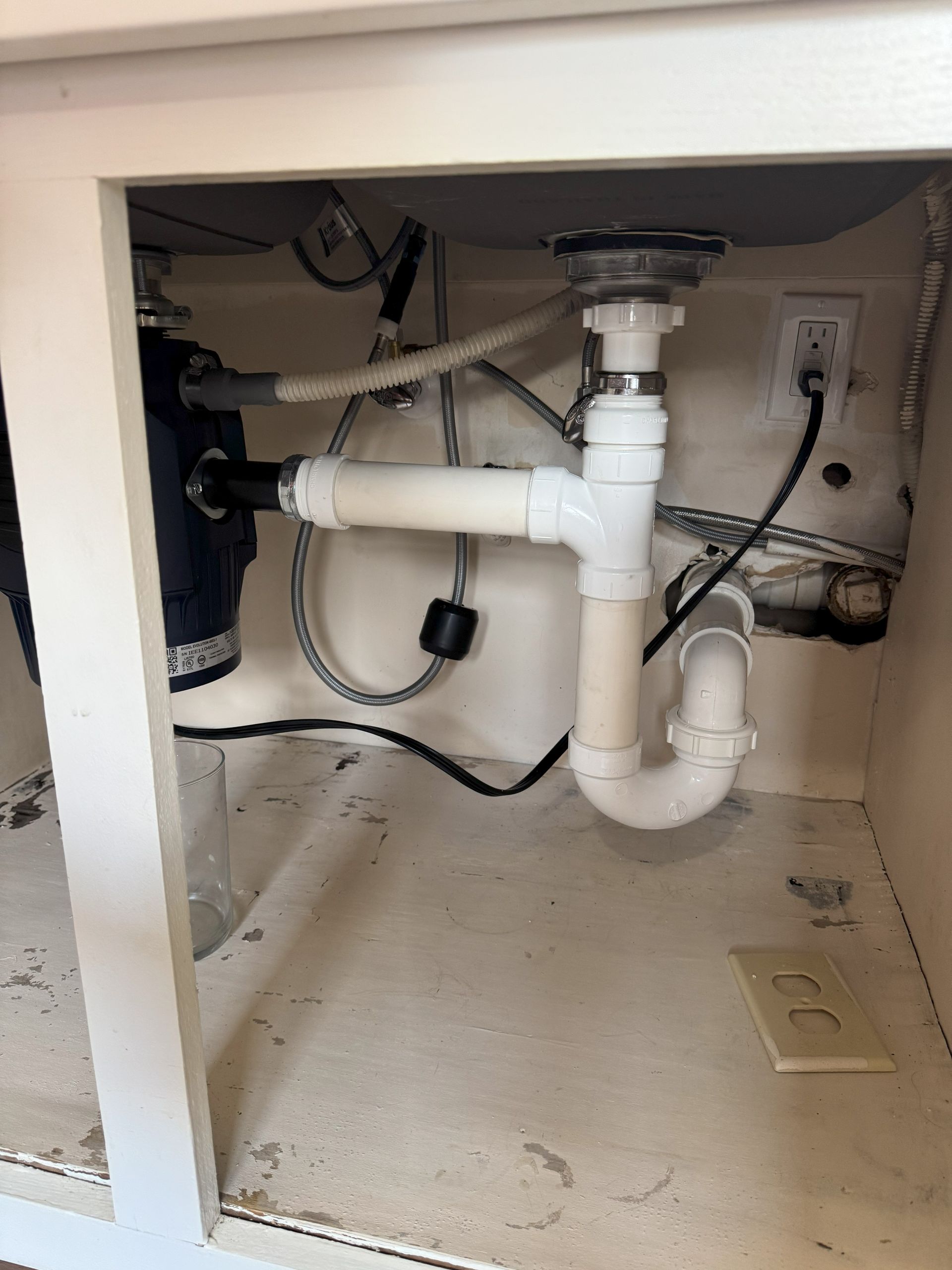 Under-sink plumbing with a garbage disposal, pipes, and an electrical outlet within a white cabinet.