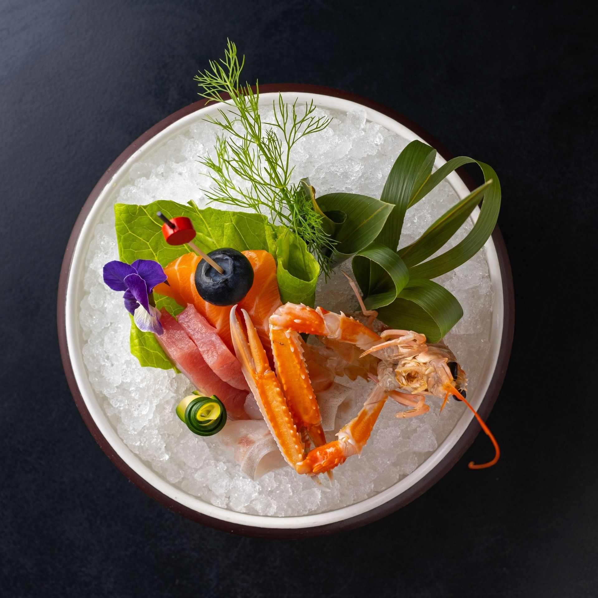 Piatto di sashimi e scampi su ghiaccio, guarnito con verdure e un mirtillo.