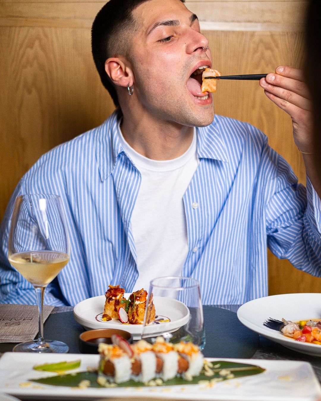 Uomo che mangia sushi con le bacchette, con camicia bianca e camicia a righe blu, e tavola apparecchiata con cibo e vino.