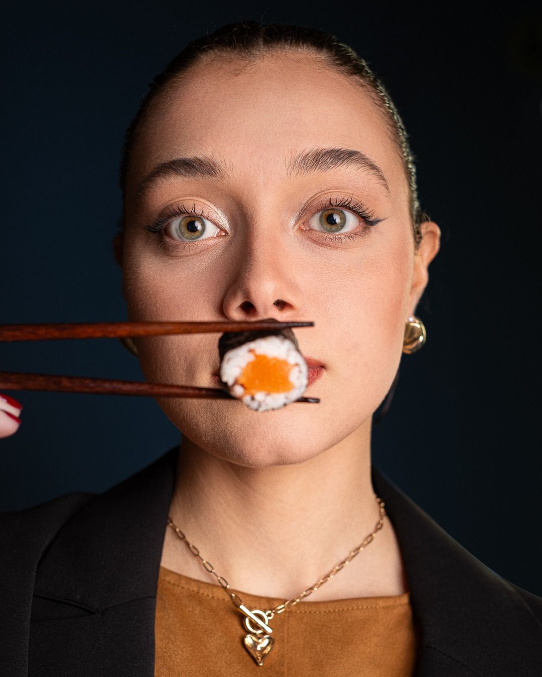 Donna che tiene in mano un rotolo di sushi con le bacchette, con aria sorpresa, su uno sfondo blu scuro.