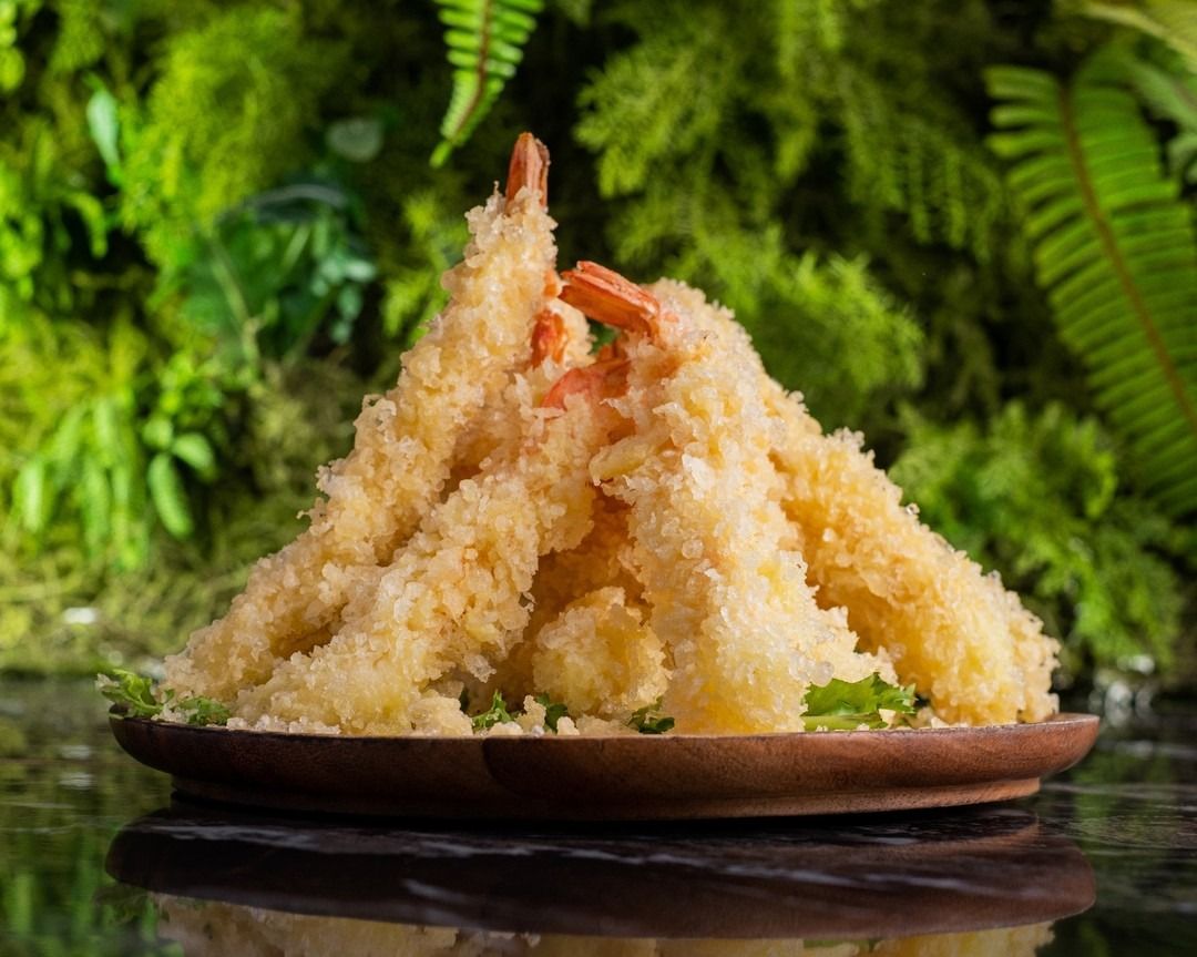 Gamberi in tempura disposti su un piatto di legno, con uno sfondo di foglie verdi.