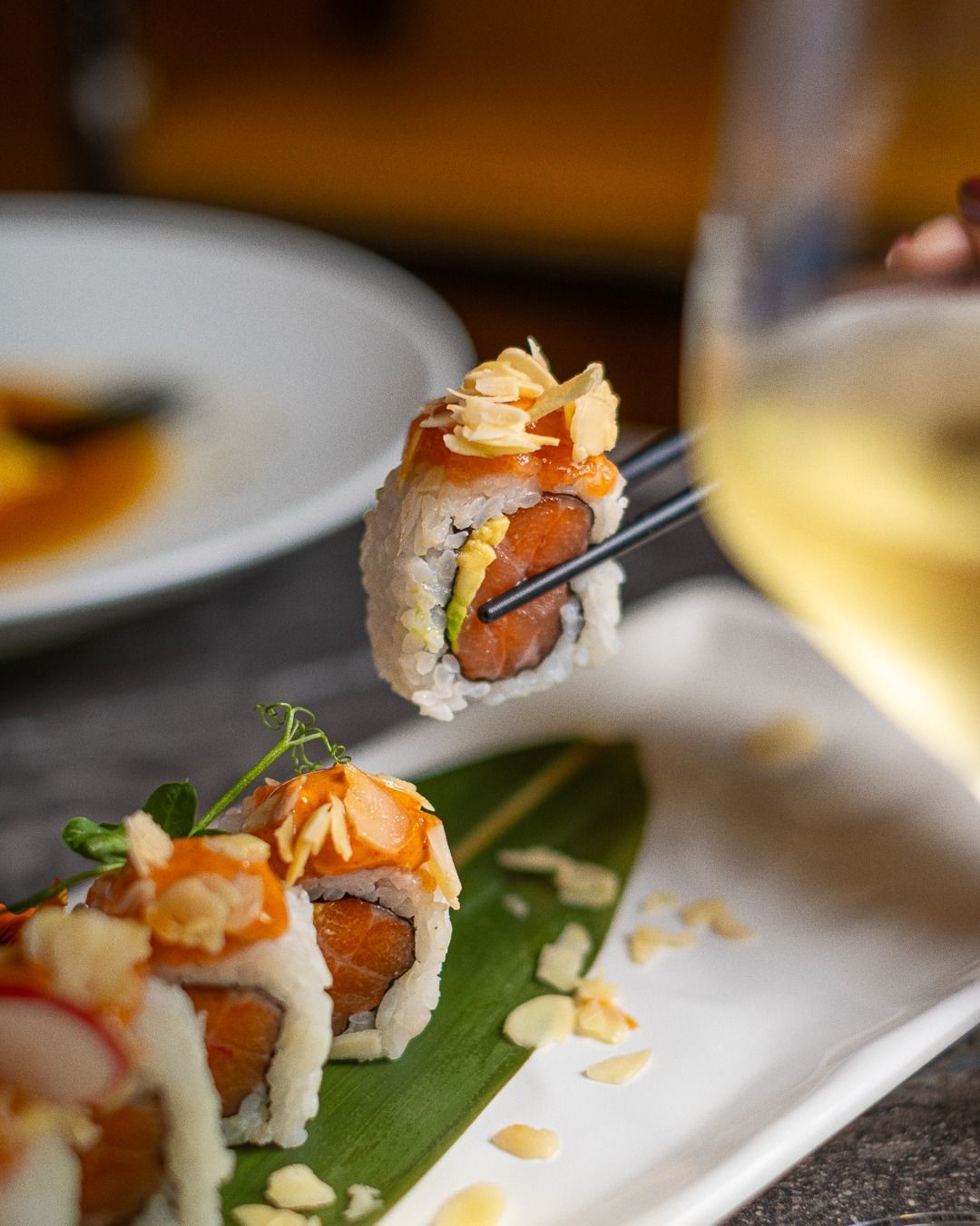 Rotolo di sushi tenuto con le bacchette, vicino a un bicchiere di vino e a un piatto.