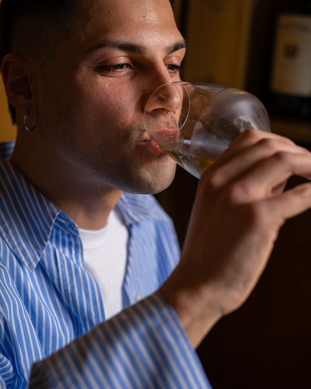 Un uomo beve da un bicchiere, indossa una camicia a righe blu e una canottiera bianca. Si trova in una stanza buia.