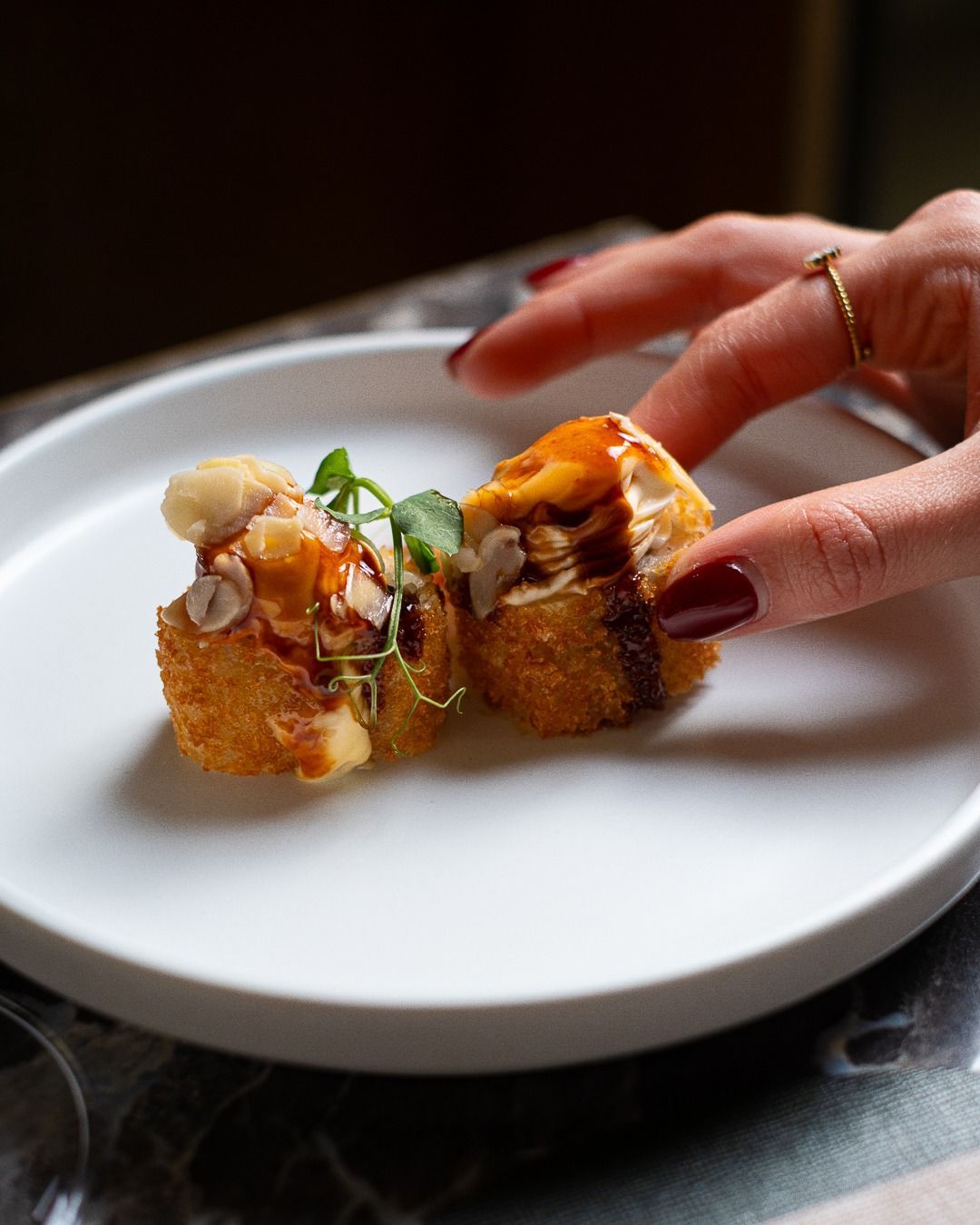 Mano che tiene un panino fritto, diviso in due e condito, su un piatto bianco.