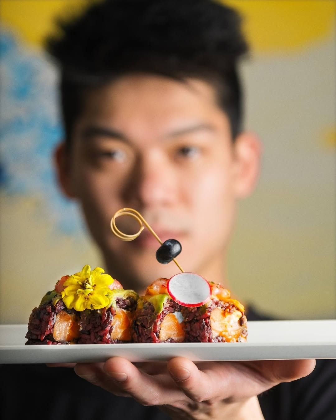 Chef che tiene in mano un piatto con piccole porzioni di cibo, guarnito con fiori e olive.