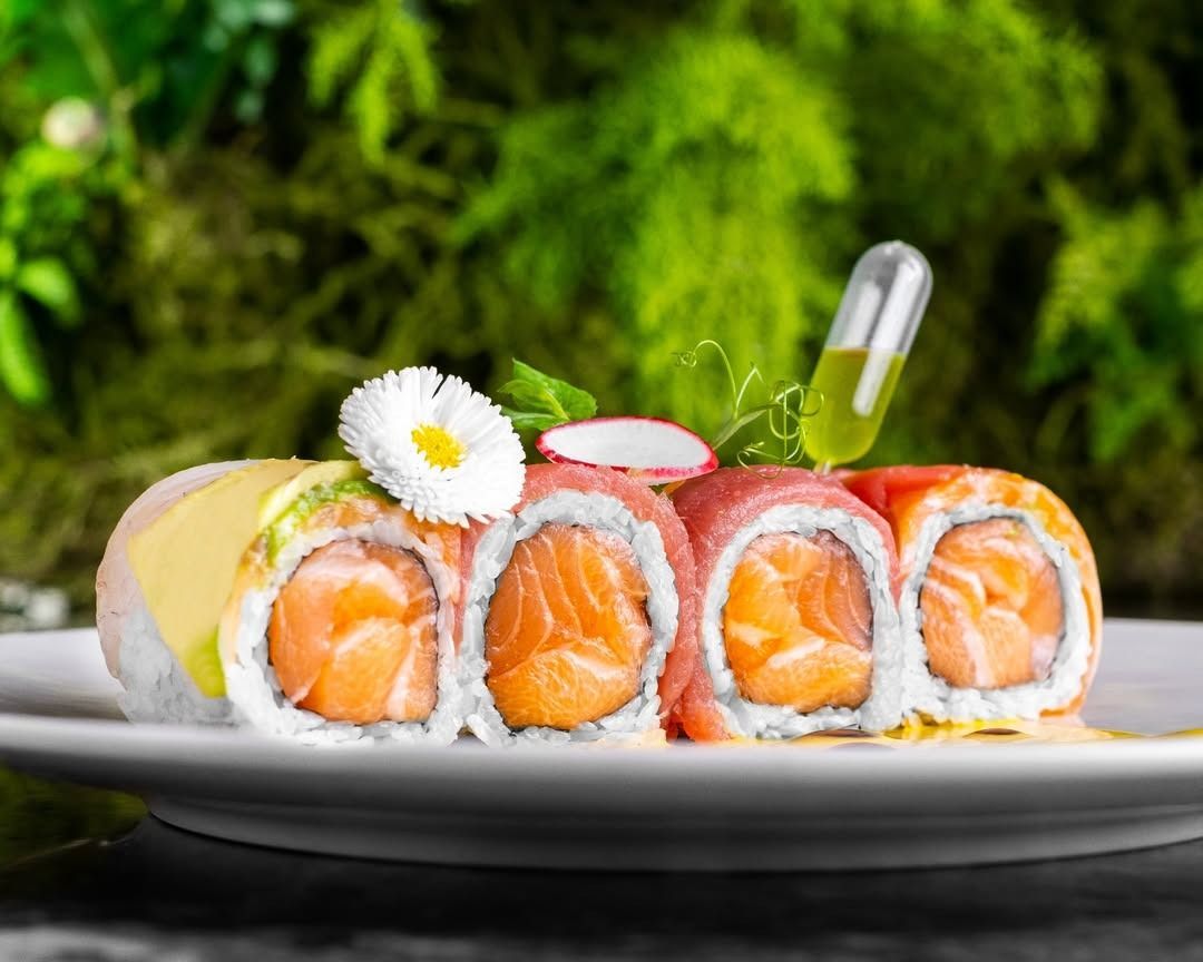 Involtini di sushi su un piatto bianco, guarnito con un fiore e un ravanello; sfondo verde sfocato.