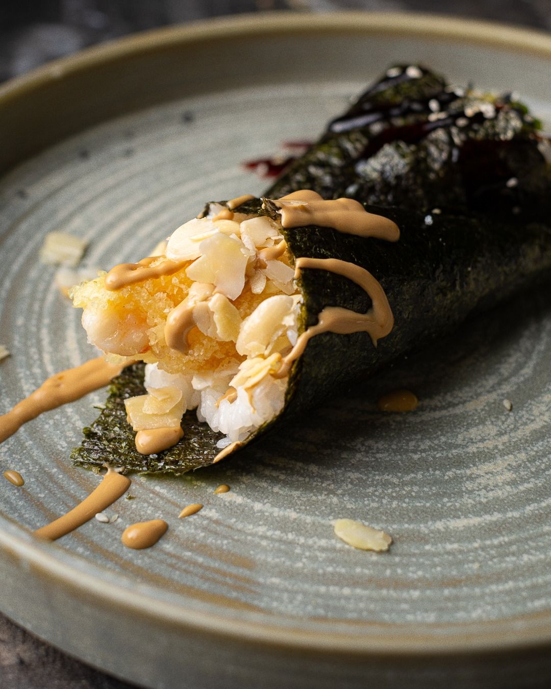 Cono di sushi Temaki, ripieno di gamberi e guarnito con salsa e mandorle, servito su un piatto grigio.