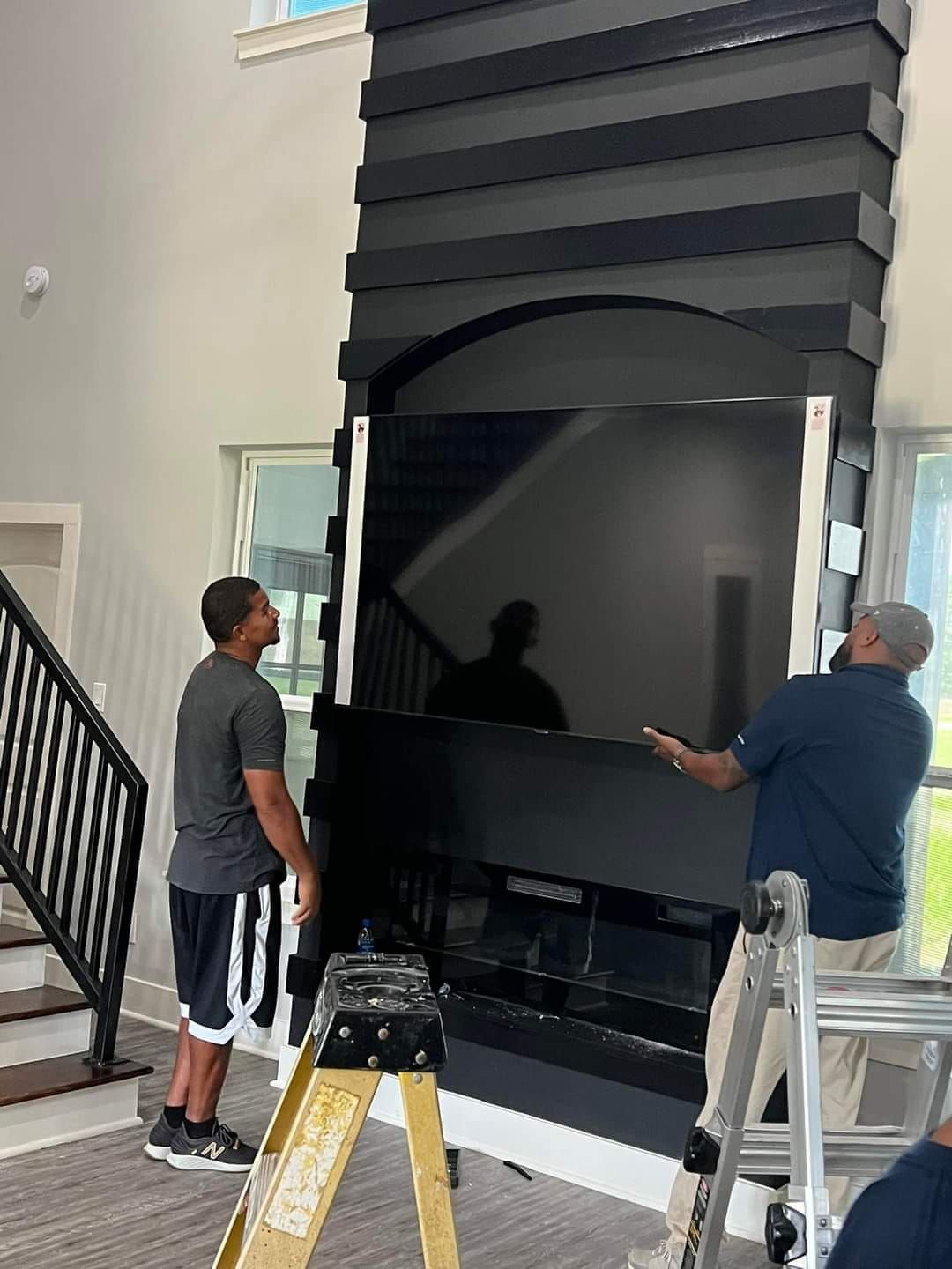 Two people installing a large TV on a black fireplace. They are standing near stairs.