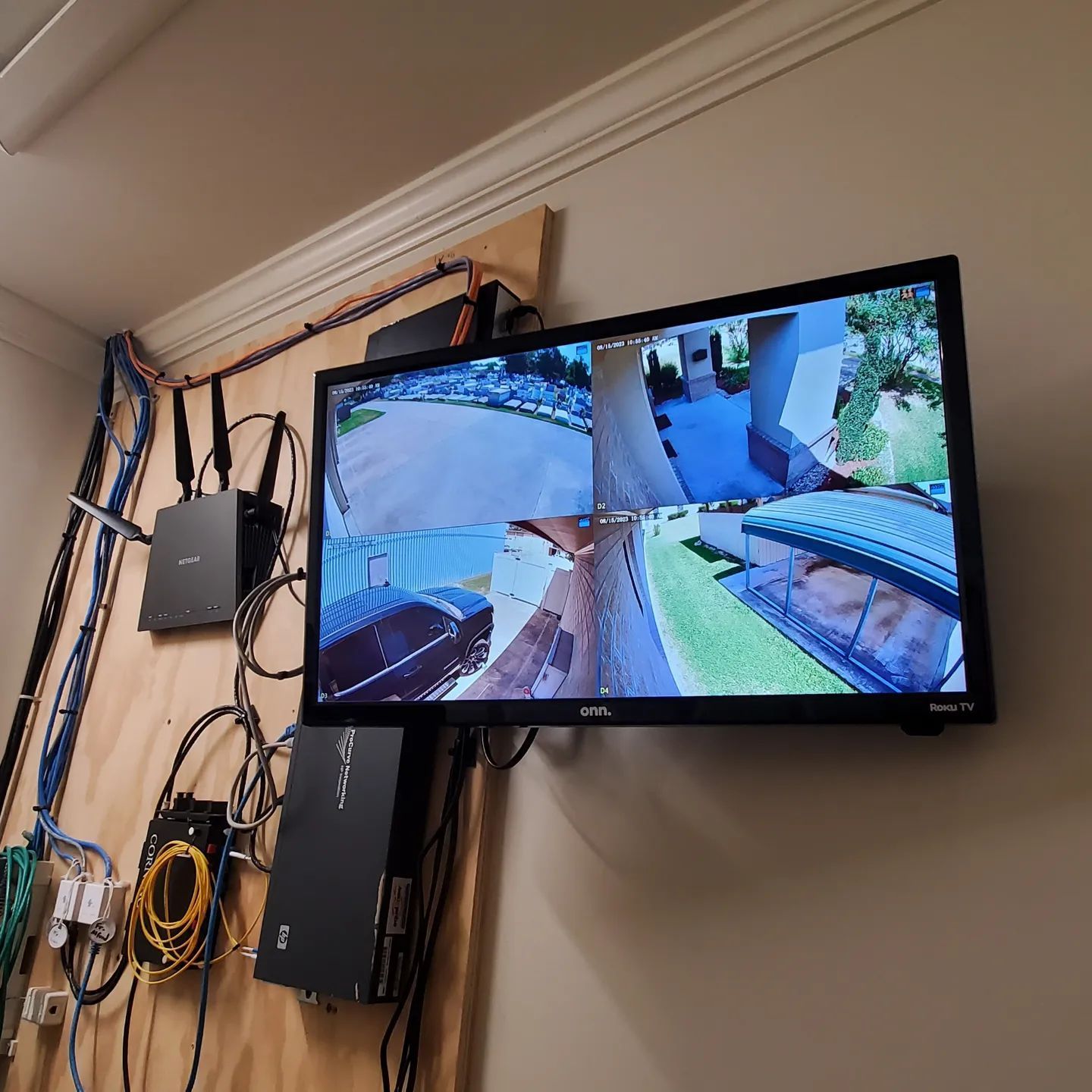 Security camera monitor showing four views, mounted on a wooden board with networking equipment.