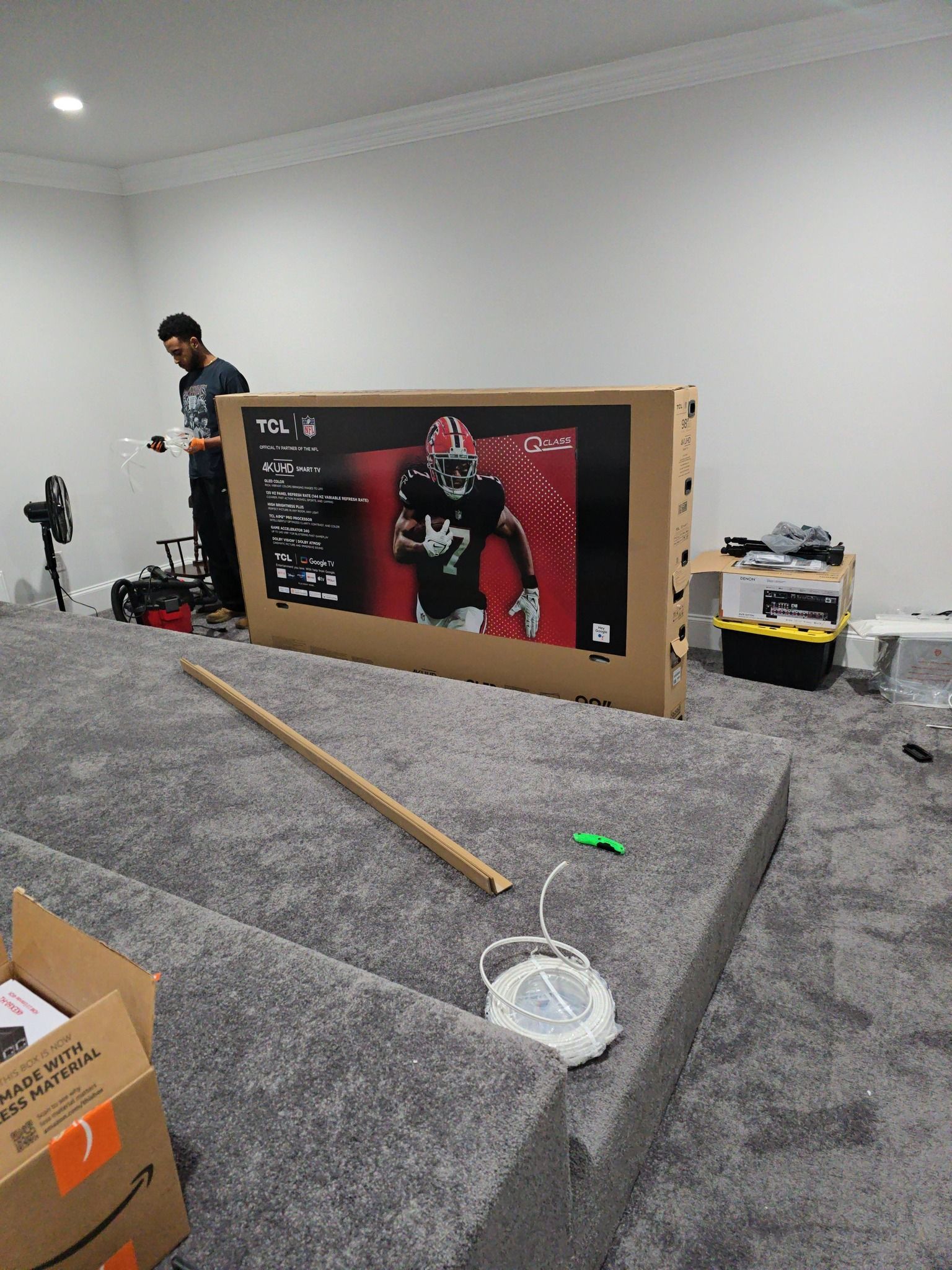 Man installing TV in a room with gray carpeted steps. Box, tools, and TV screen showing a football player are visible.
