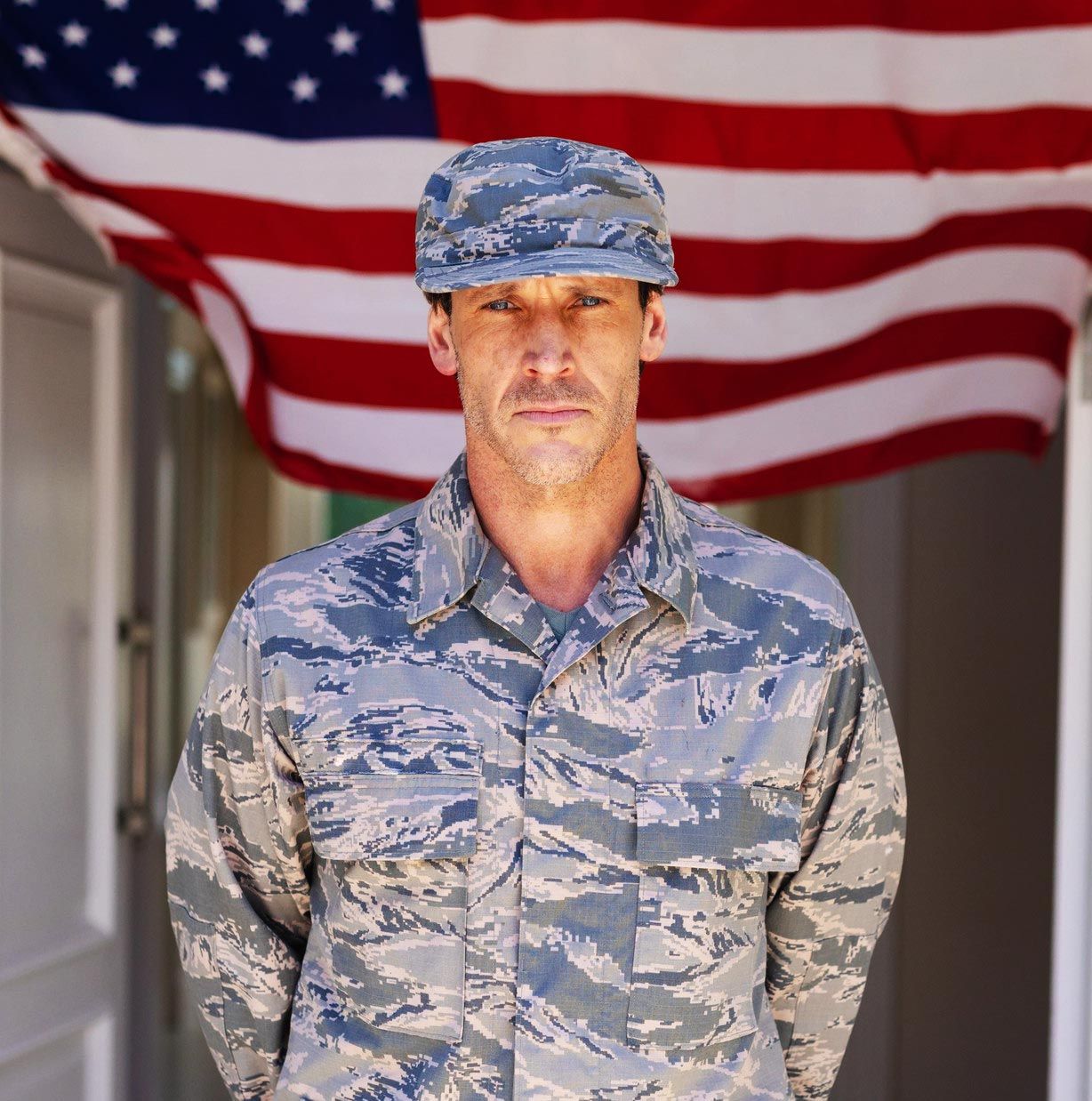 A person in Air Force digital camouflage uniform and cap stands centered in front of an American flag.