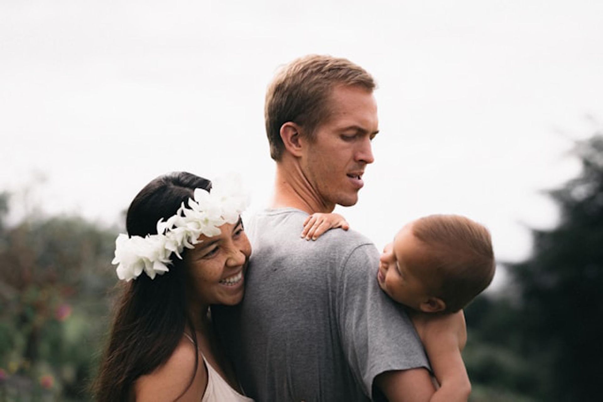 A person with a white flower crown smiles while resting their head on the shoulder of another person holding a child.