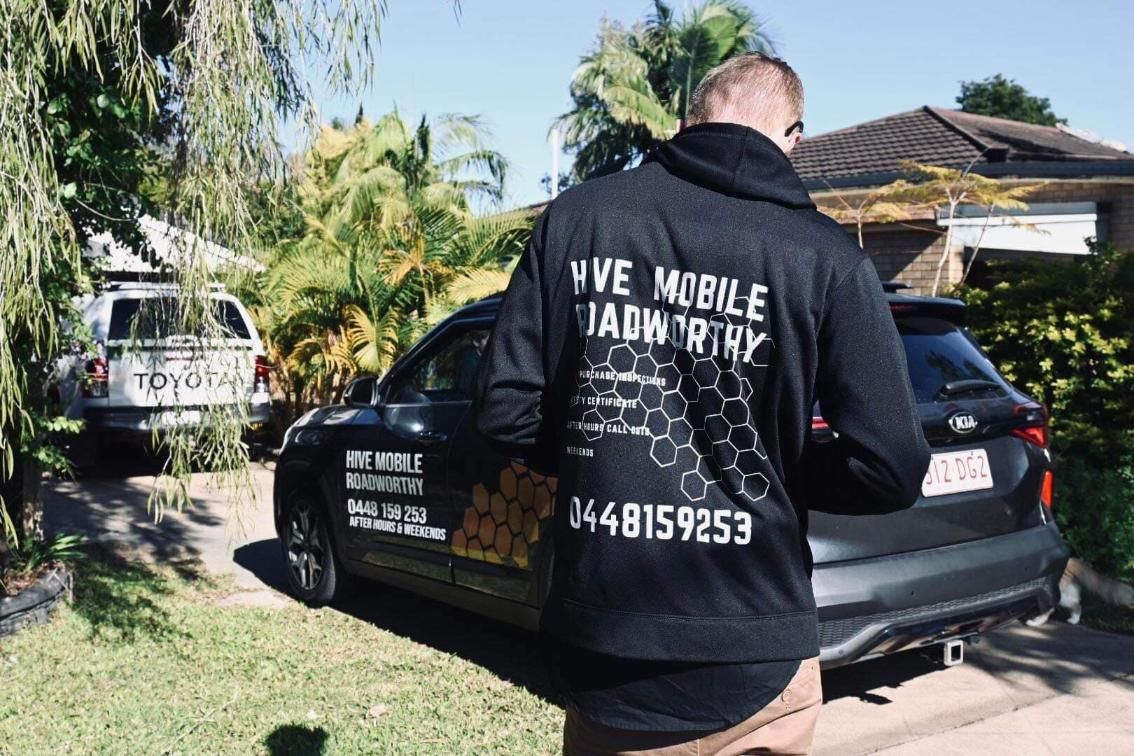 A Man Wearing a Black Hoodie is Standing in Front of a Car — Hive Mobile Roadworthy in Highland Park, QLD