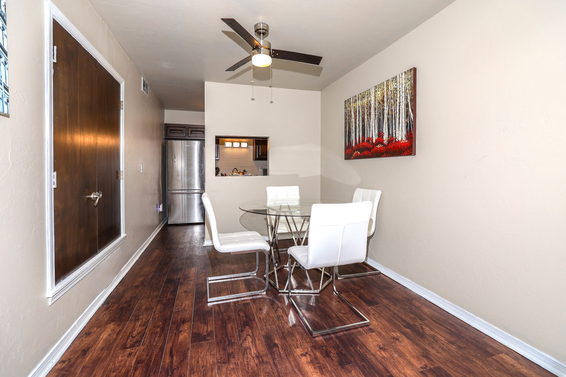 Dining area with glass table and white chairs, dark wood floor, and painting on wall.