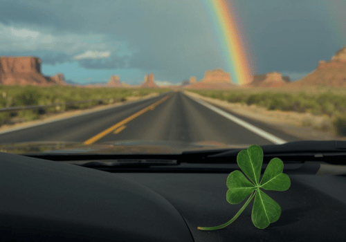 Dashboard view of a desert highway with a rainbow ahead and a four-leaf clover on the dash.