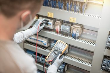 Ein Mann mit Maske und Handschuhen verwendet ein Multimeter in einem Schaltschrank.