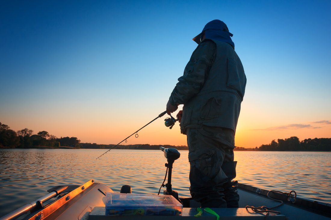 A man is fishing in a boat on a lake at sunset.