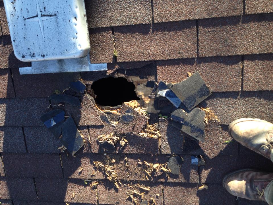 Hole in roof shingles next to a downspout. Shingles are broken and torn. A person's feet are visible.