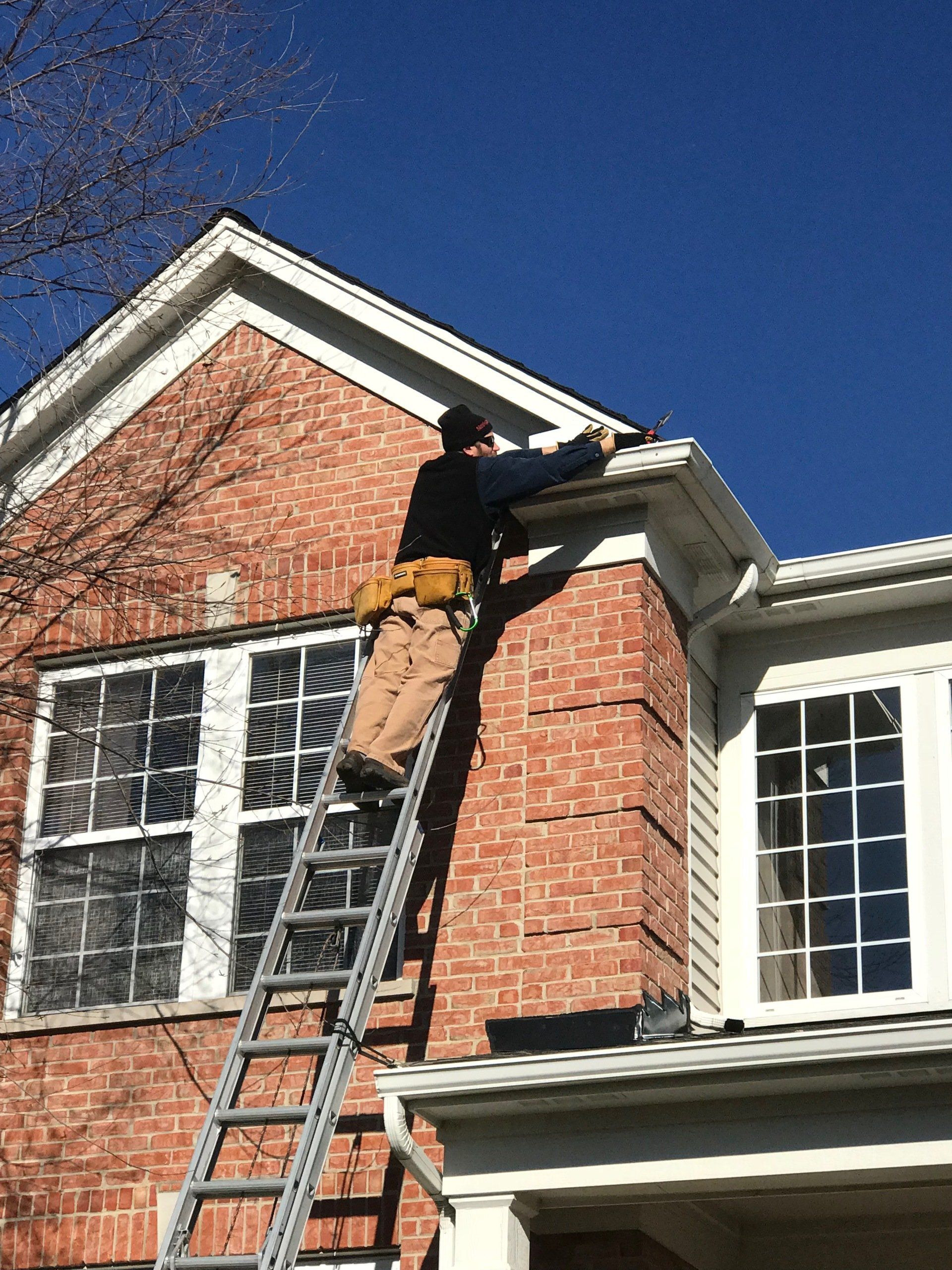 A person on a ladder is working on the gutter of a brick building on a sunny day.