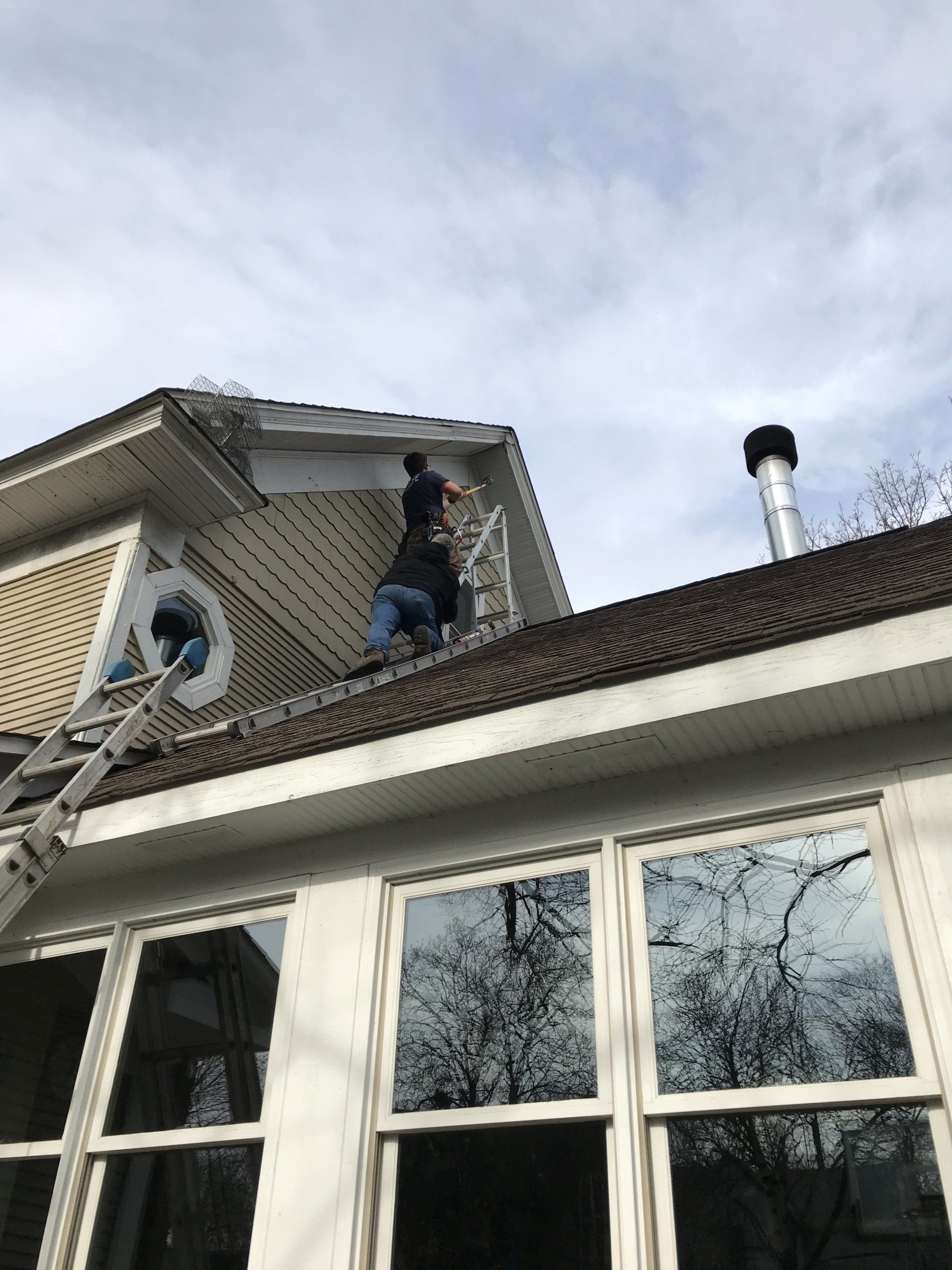 Person on a ladder repairs the roof of a house with windows and a chimney under a cloudy sky.