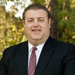 A professional headshot of a person wearing a dark pinstripe suit, white collared shirt, and a patterned red tie.