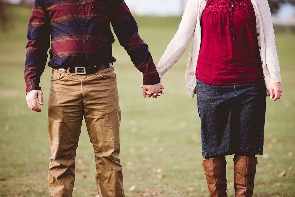 A couple holds hands while standing in a grassy field, wearing casual fall clothing including boots and a flannel shirt.
