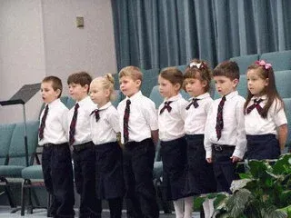 A line of children in white shirts, dark ties, and dark skirts or pants standing in a row on a stage.