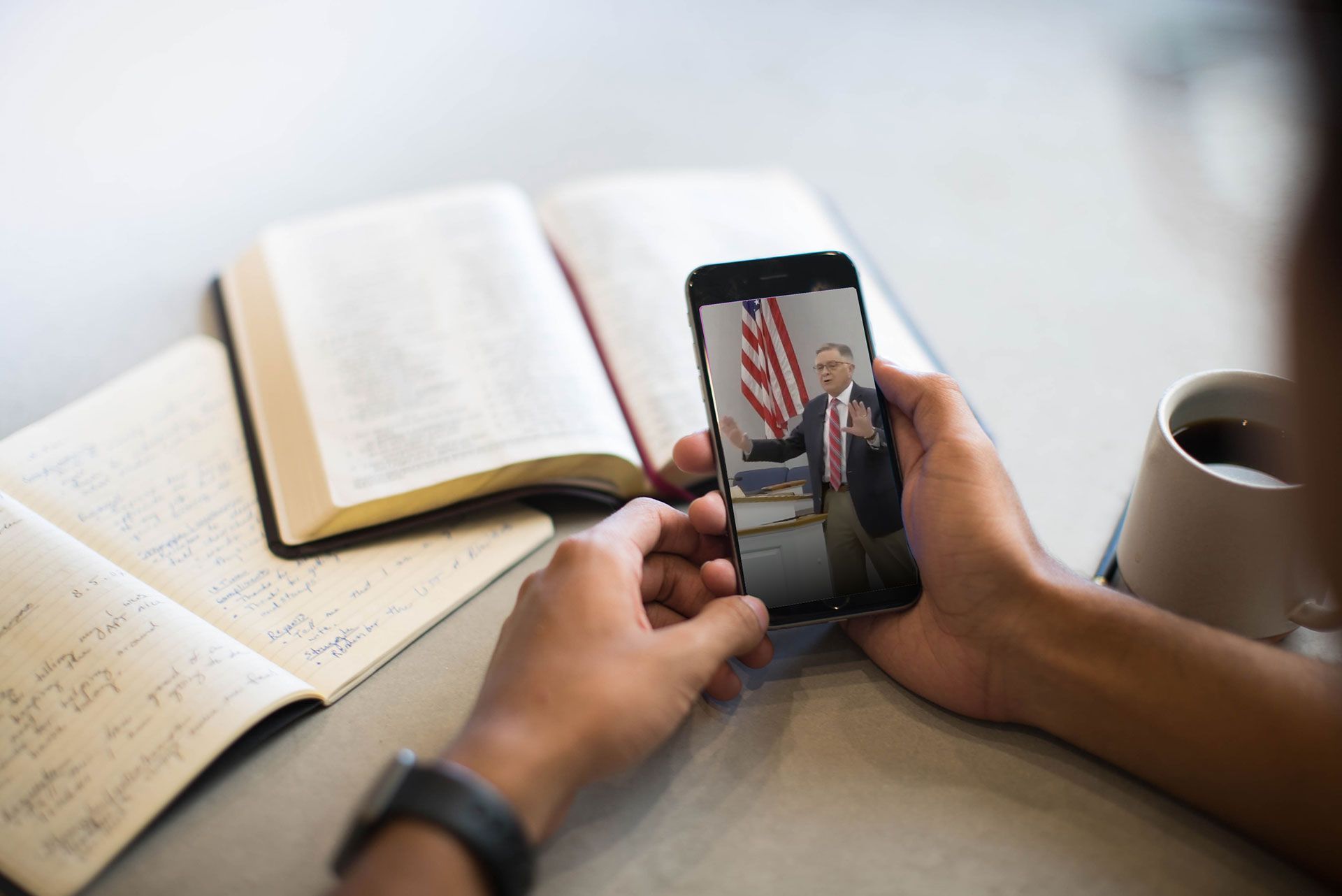 Hands hold a smartphone displaying a video of a person standing before an American flag, next to a notebook and a mug.