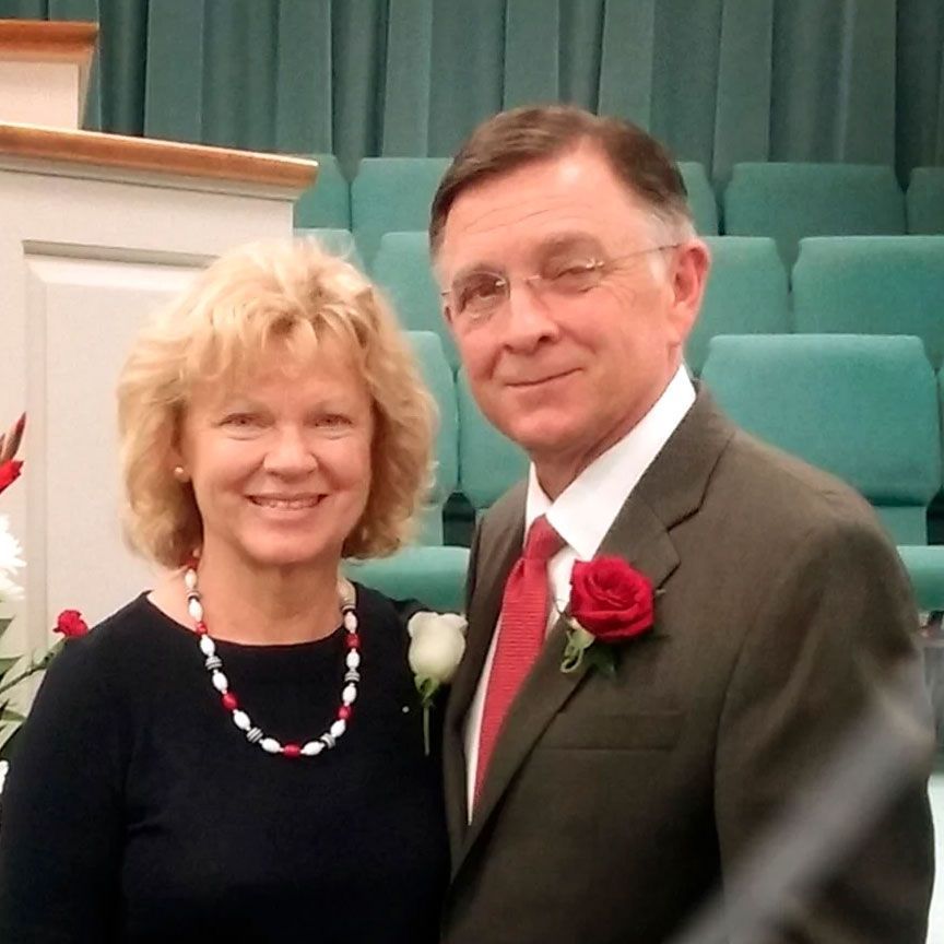 A smiling couple stands together indoors in front of teal chairs and a podium, wearing a rose boutonniere and a necklace.
