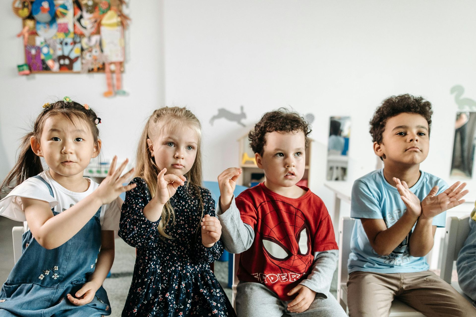 Four children sitting side by side, engaging in a classroom activity with expressive gestures and focused expressions.