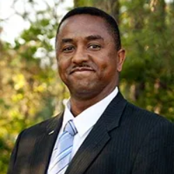 A man in a black pinstriped suit, white shirt, and blue-striped tie smiles outdoors against a blurred foliage background.