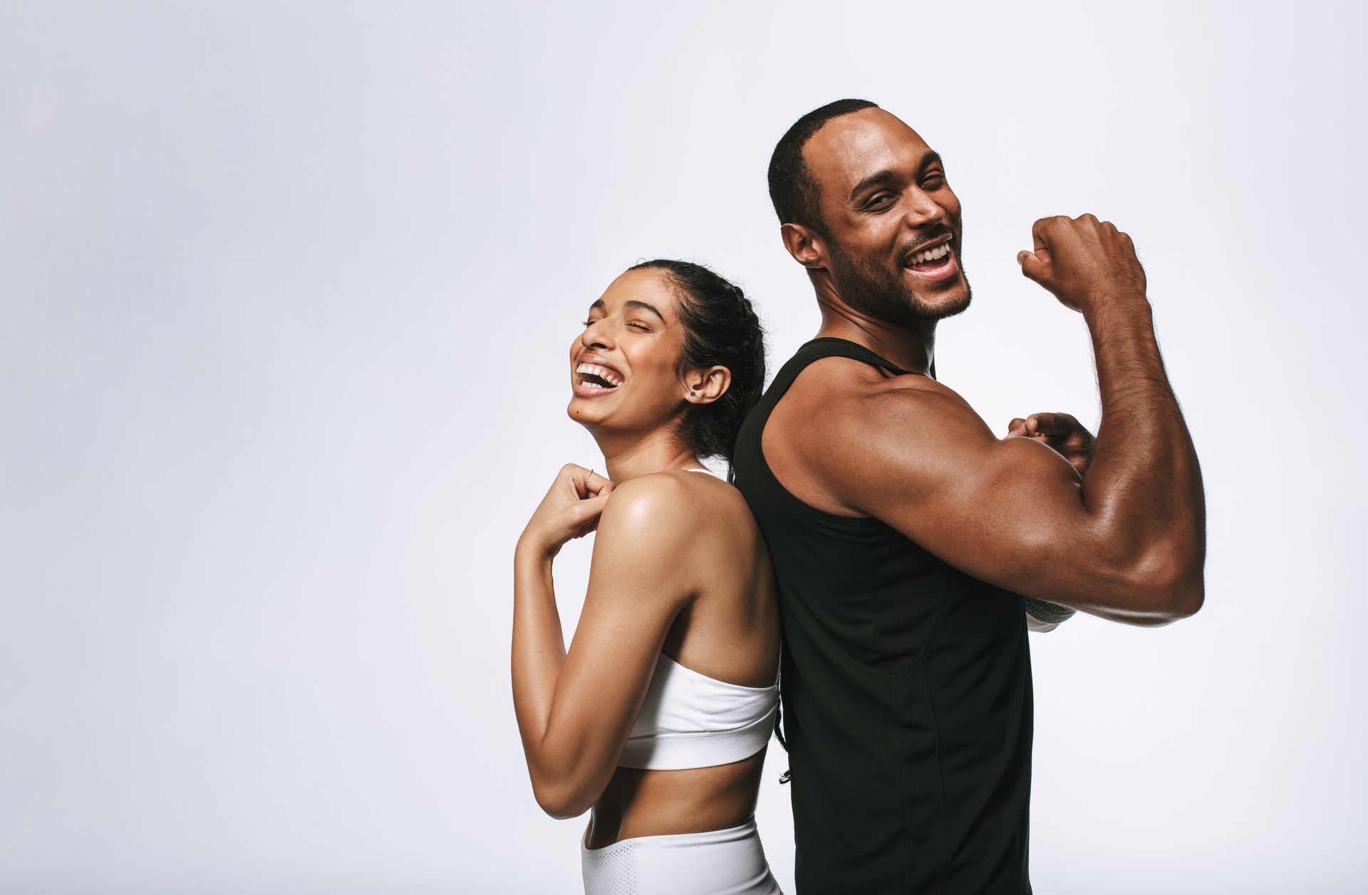 Smiling people back-to-back, flexing muscles. One in white sports bra, other in black tank top.