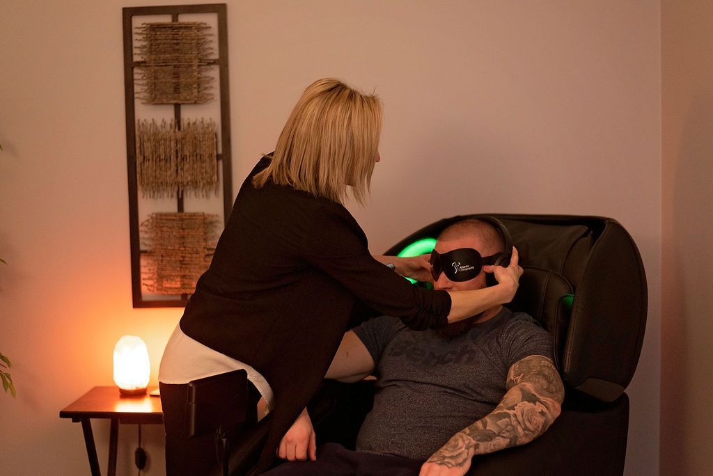 Woman placing eye mask on a person seated in a chair, likely a relaxation or therapy setting.