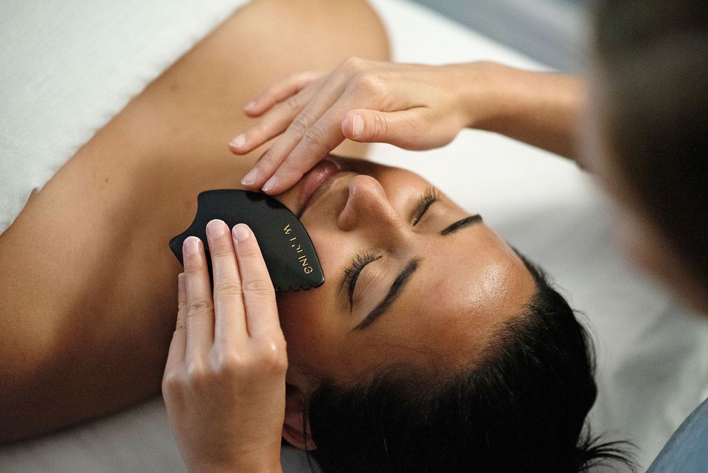 Person receiving facial gua sha treatment with black tool, eyes closed on a massage table.