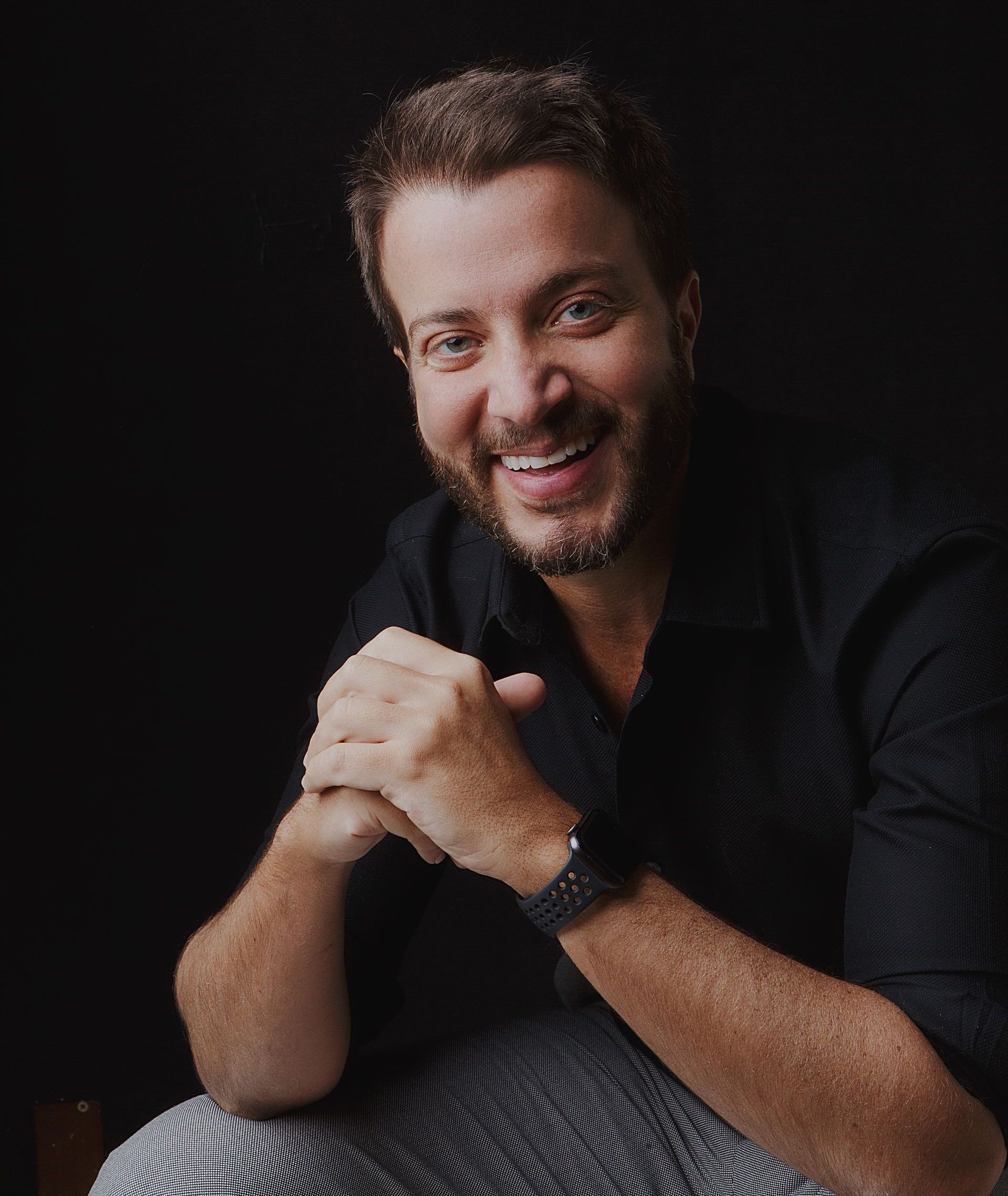 Homem com cabelo castanho curto e barba, sorrindo, vestindo uma camisa preta, mãos entrelaçadas, fundo escuro.