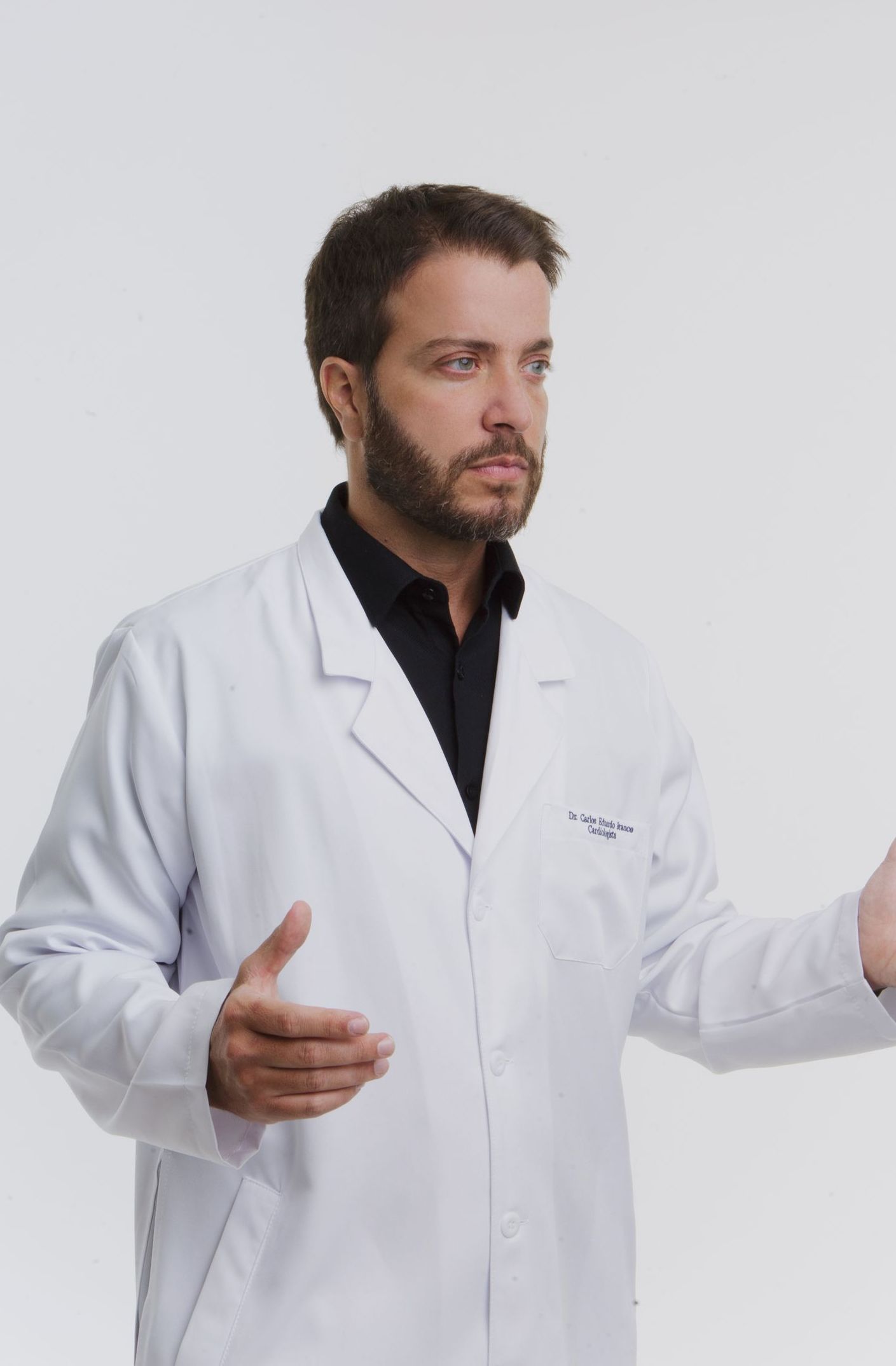 Homem de jaleco branco, mãos abertas, camisa escura, olhando para longe, cenário de estúdio.