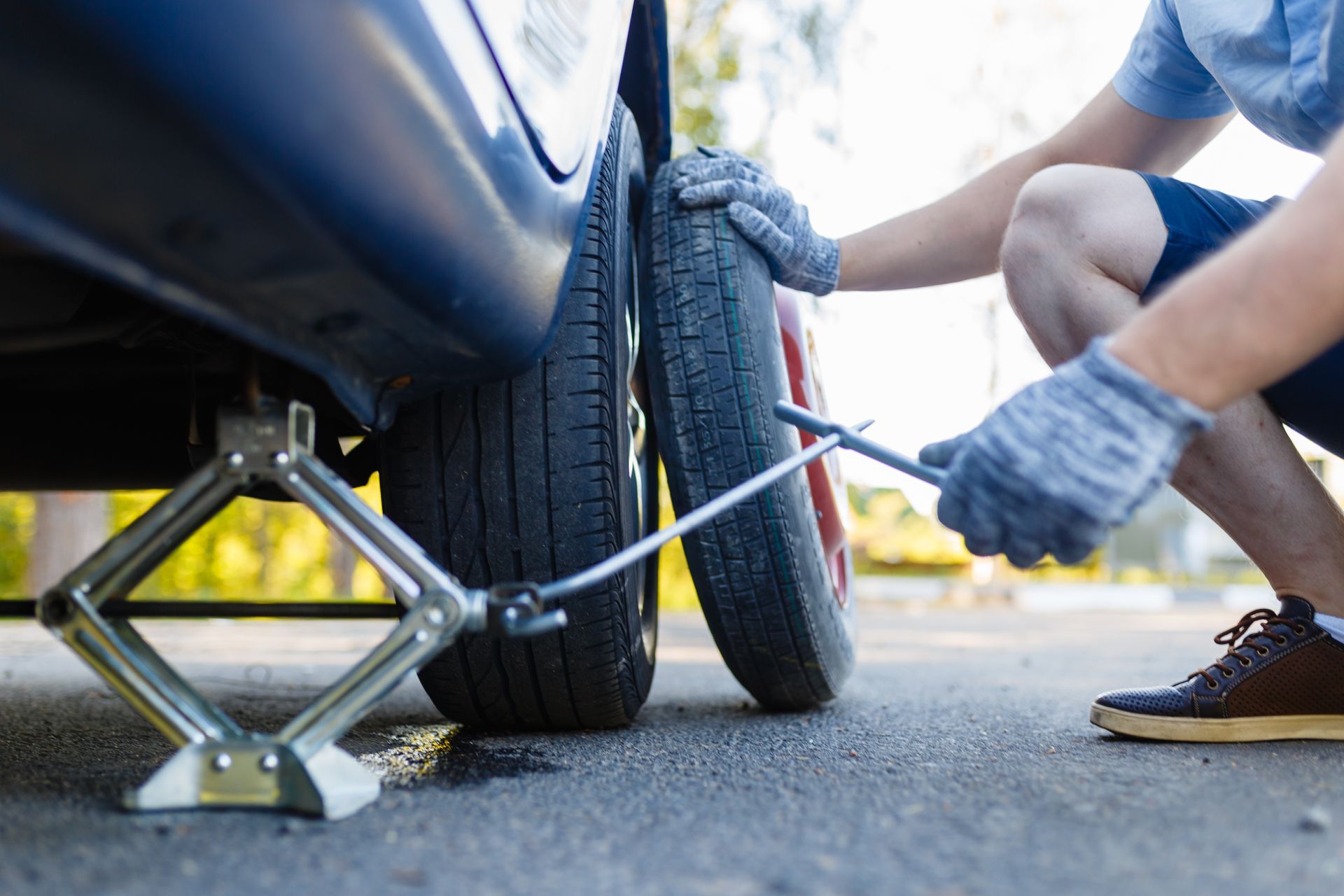 A person wearing gloves using a metal jack to lift a blue car to change a flat tire on a paved road.