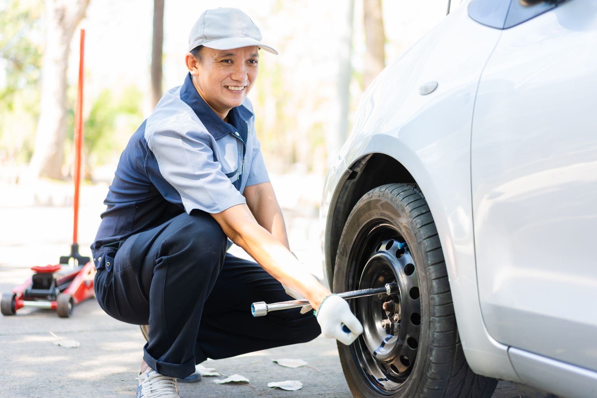 A technician in uniform uses a lug wrench to tighten the wheel nuts on a silver car, with a floor jack visible nearby.