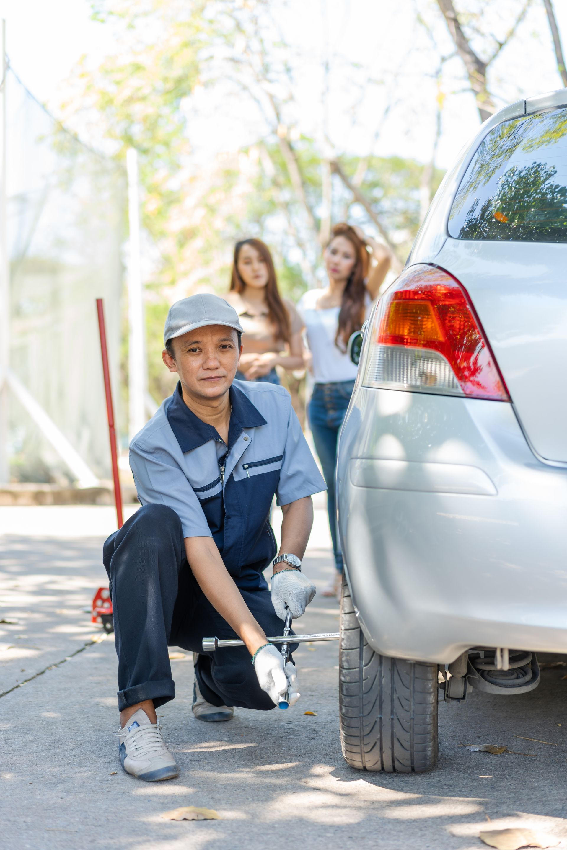 A mechanic in a blue uniform crouches by a car tire with a tool, while two individuals stand in the blurred background.