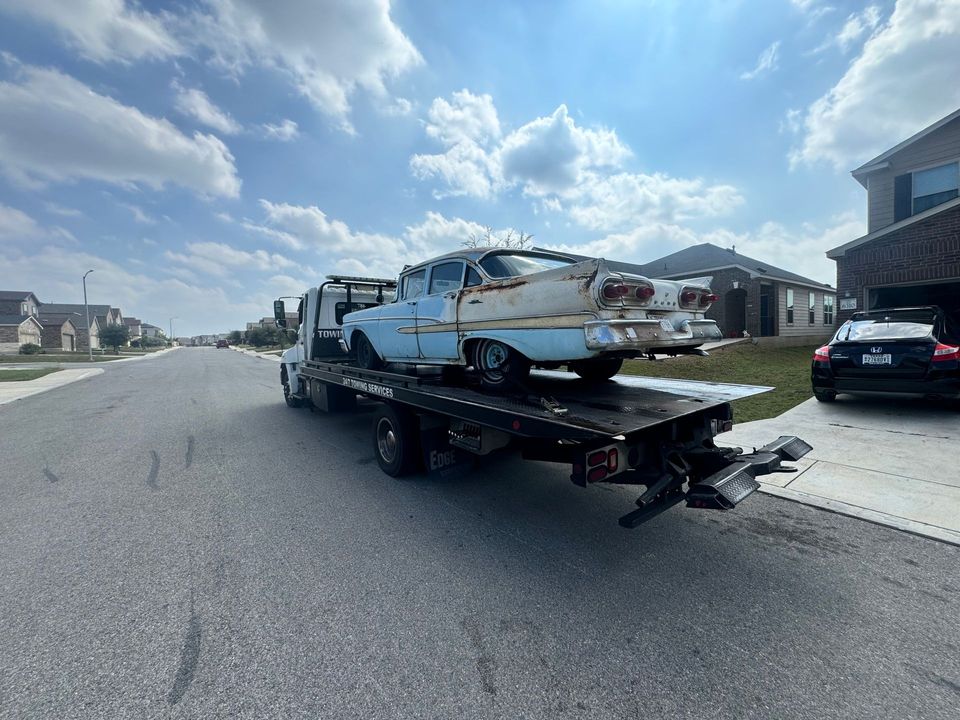 A light blue, weathered vintage car being transported on a flatbed tow truck on a suburban residential street.
