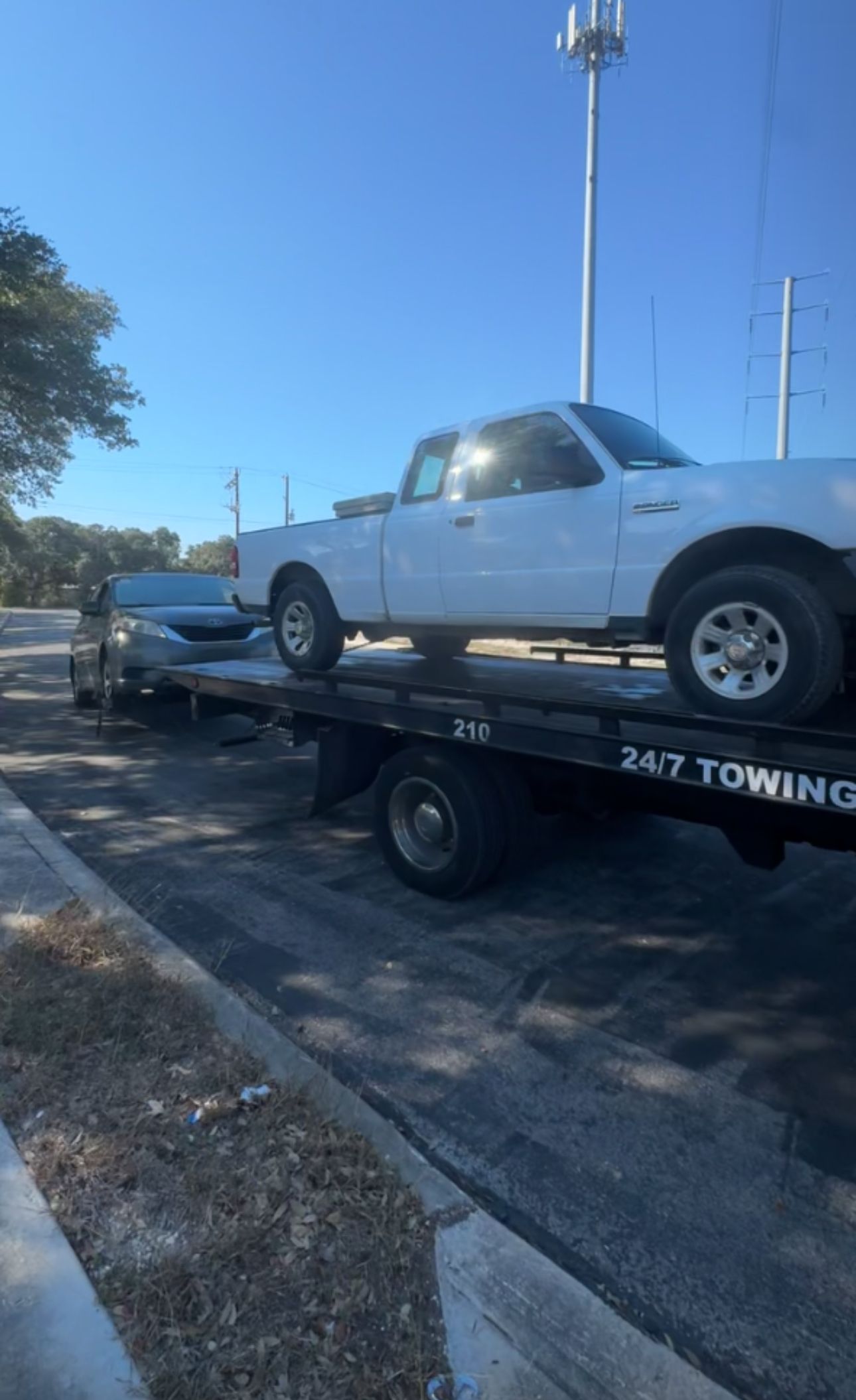 A white pickup truck sits on the back of a flatbed tow truck parked outdoors under a bright blue sky.