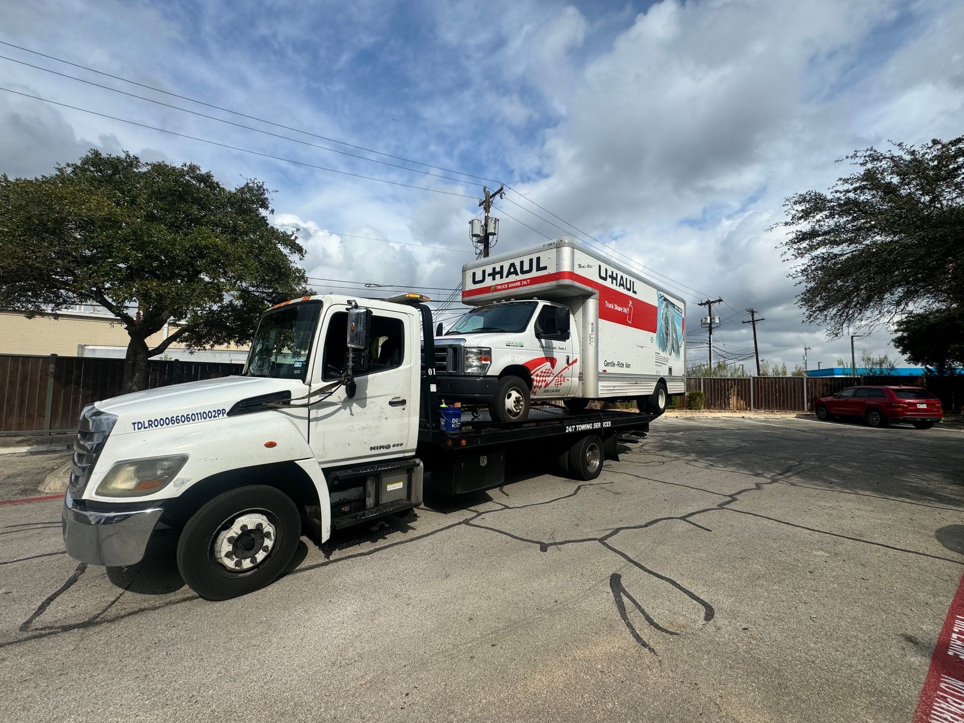 A white tow truck hauling a U-Haul box truck in a parking lot under a cloudy sky.