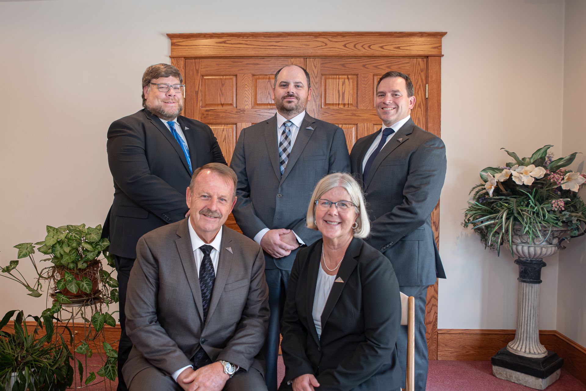 Five people in suits, posed in a room with a wooden door and a large vase of flowers.