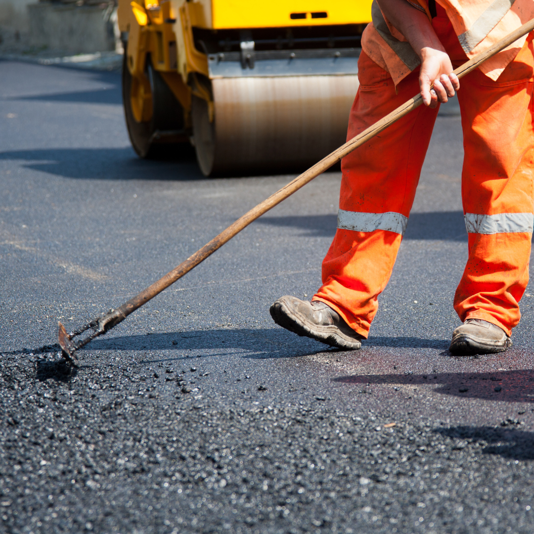A man in orange pants is using a paver to spread asphalt on a road