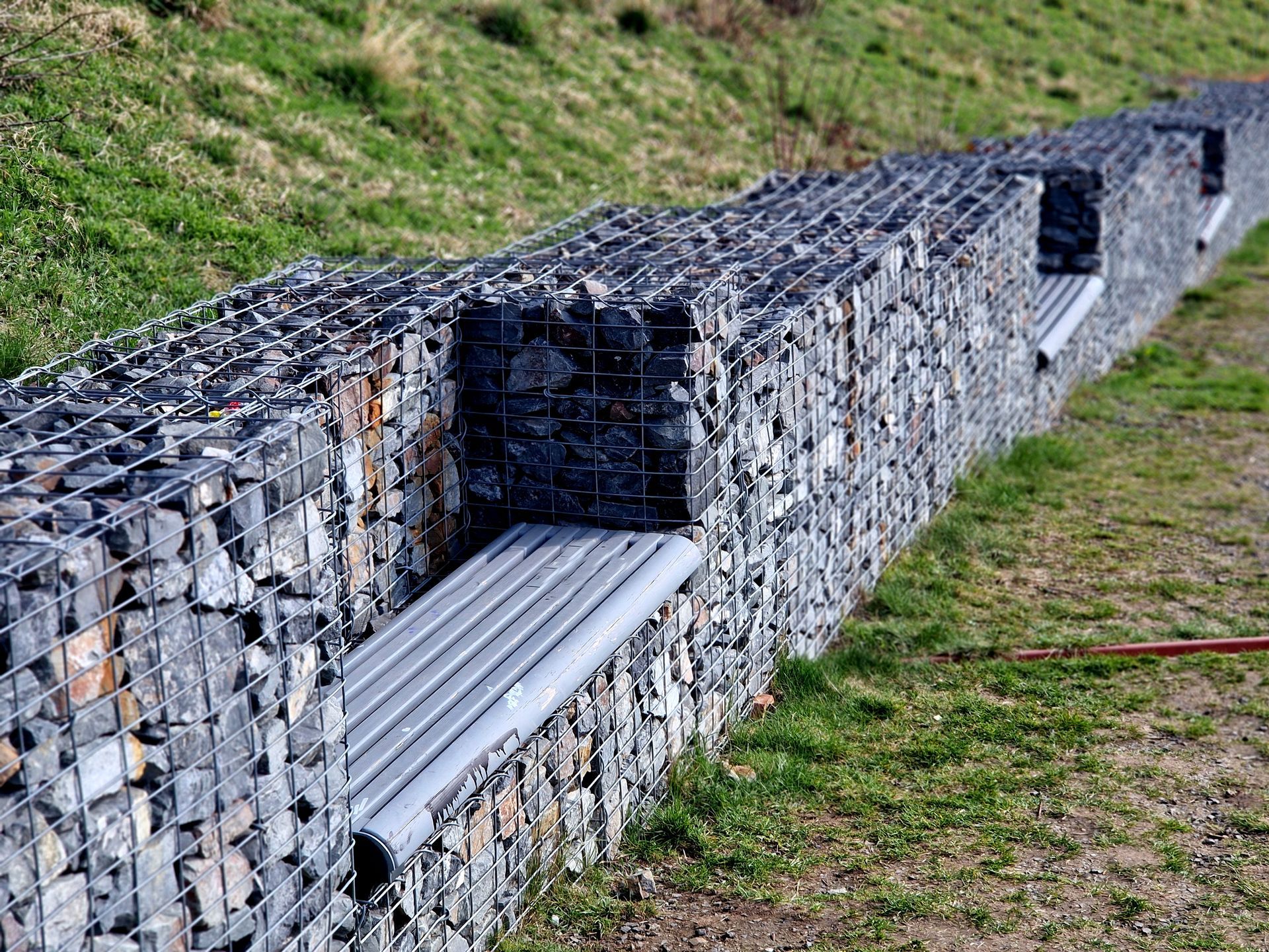 Gabion retaining walls with pipes for drainage and wooden benches inserted, built into a dry wall.