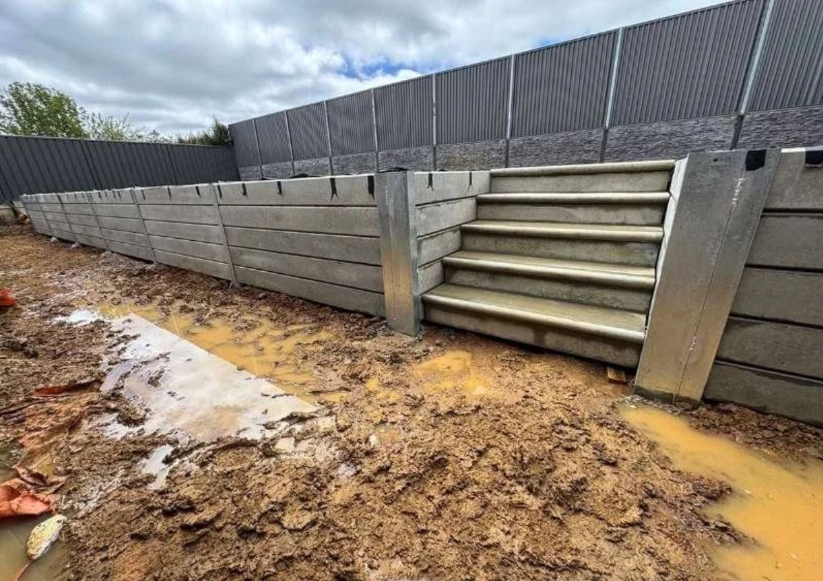 Muddy flooded yard beside concrete retaining wall and steps under overcast sky
