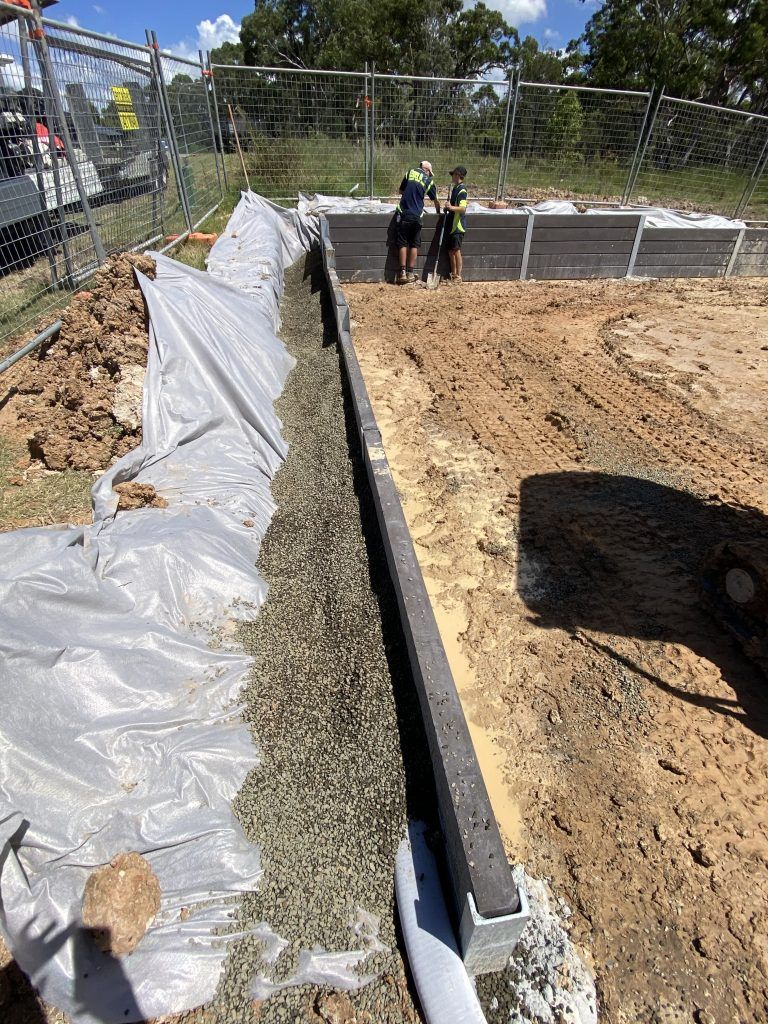 Construction site trench lined with white plastic, gravel channel, and workers in the background